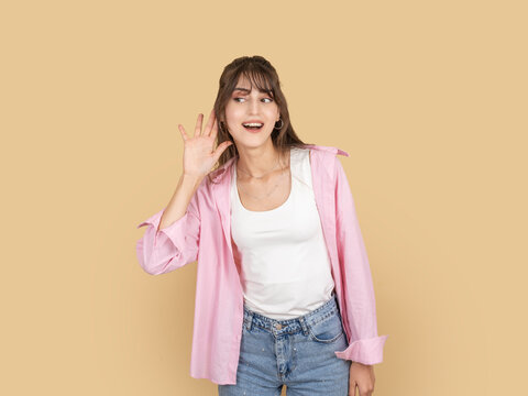 Curious young woman cupping hand to ear and smiling, trying to hear or listen closely concept idea image. Isolated on beige background with copy space for advertising and marketing use.