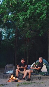 Relaxing outdoors at sunset. Two individuals resting beside campfire. Peaceful evening with friends camping in nature. Vertical video. Two men savor moment near their tent during evening in woods.