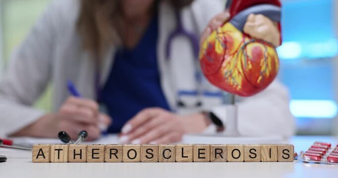 Wooden blocks spell word Atherosclerosis near heart model and tablets. Doctor writes chart reviewing cholesterol risks while updating care plan