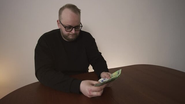 Man exhales in disappointment while counting euro banknotes at table. Concept of not enough money, financial stress, unmet needs and budgeting struggle.