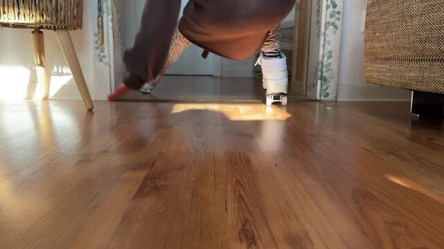 Low-angle view of a child roller skating indoors and falling in a funny, playful moment. Captures humor, movement, and candid childhood activity on a wooden floor in warm home lighting.