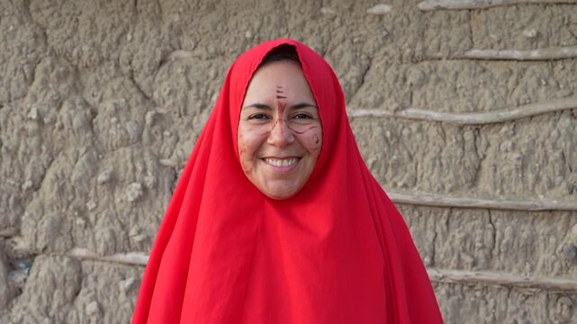 Portrait of a smiling Wayuu woman in traditional red shawl with facial markings, standing by a rustic adobe wall in La Guajira, Colombia. Soft daylight, static close-up shot.