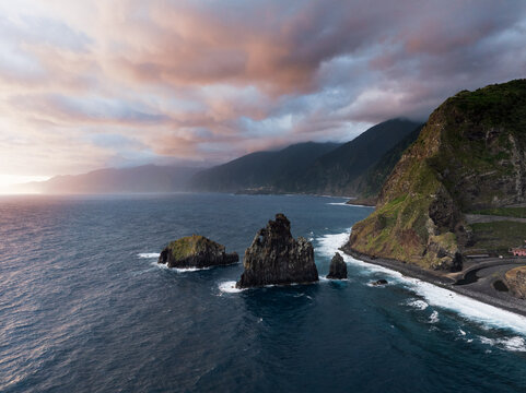 Aerial view of Ribeira da Janela sea stacks rising from the ocean near steep green cliffs under a dramatic sunset sky in Ribeira da Janela, Madeira, Portugal.