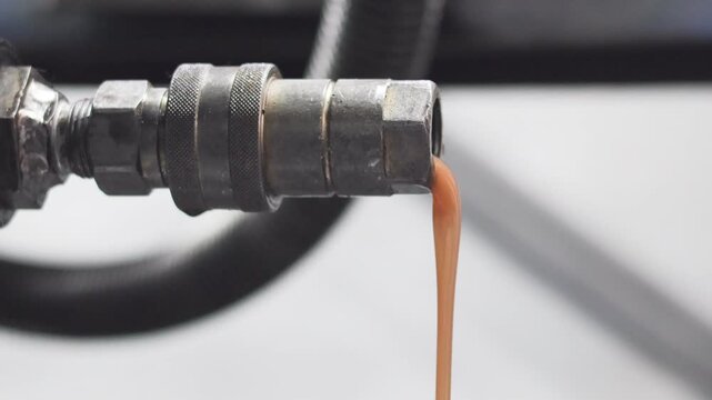 Close up of milky brown contaminated hydraulic oil draining from a metal quick release coupler. Indicates water contamination or emulsion in a heavy machinery fluid system.