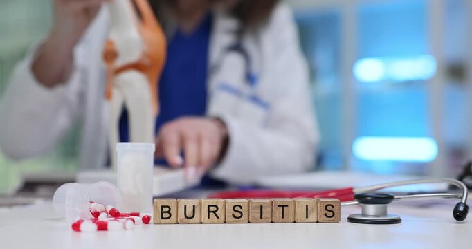Wooden blocks spell word Bursitis near joint model and stethoscope. Doctor examines chart pointing to inflamed bursa while planning rehab exercises