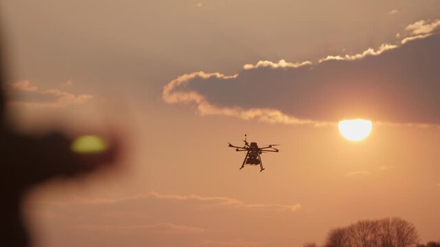 Silhouette of a drone operator's hand with a remote control, controlling a special military hexacopter, which is equipped with three explosive mines, against the background of a picturesque sunset
