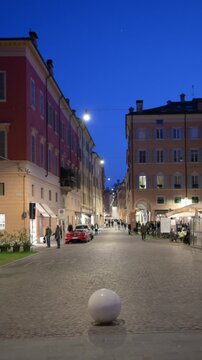Modena, italy, april 11, 2026. Evening view of a historic street in modena, italy, with people strolling past restaurants and illuminated buildings under a deep blue sky
