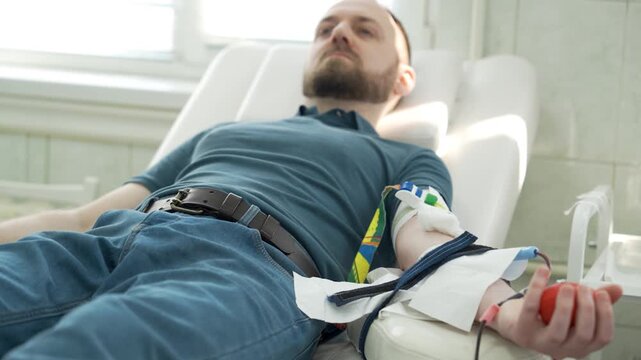 Male Donor Sitting in a Medical Chair During Blood Donation Process, Man Squeezing Red Stress Ball to Improve Blood Flow in a Modern Hospital Clinic.