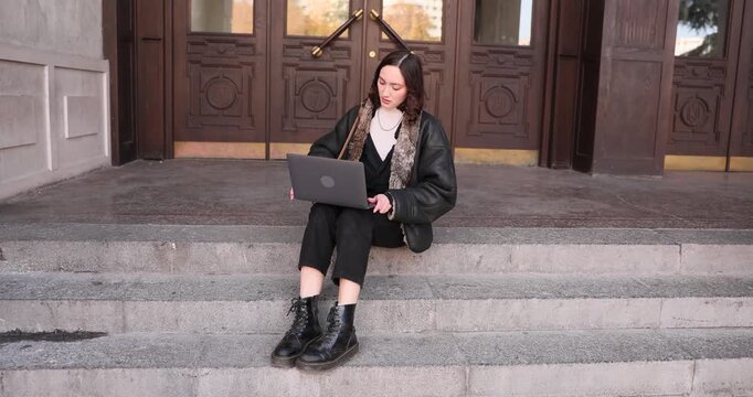 Woman student opens laptop while sits on stone steps near university building. Young female studies online by portable computer on street slow motion