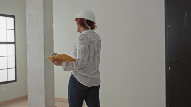 Woman architect wearing hardhat holds phone to ear while holding envelope folder in building windowed room; project planning focused.