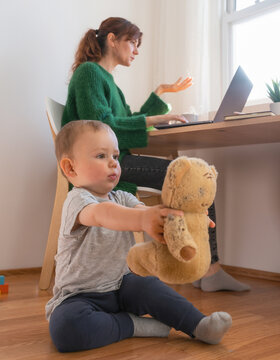 Low angle selective focus of a toddler holding a teddy bear, with a busy mother working on a laptop in the blurred background, natural window light