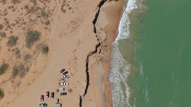 Drone video captures sweeping aerial footage of Cabo de la Vela's coastline in Colombia, showcasing striking sandy desert, turquoise waves, parked vehicles, rugged cliffs, and sunlit natural textures.