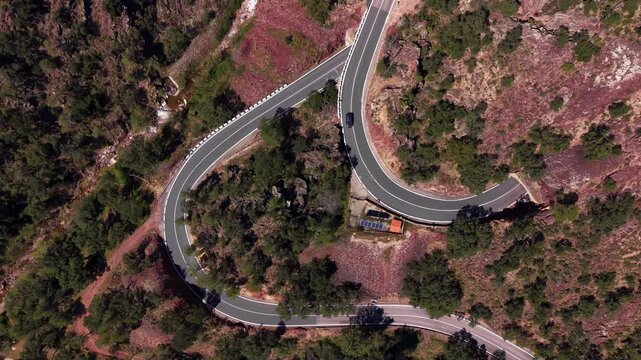 Overhead view of a winding hairpin road with a tunnel entrance carved into rocky mountainside, surrounded by sparse trees and reddish terrain with a white car visible