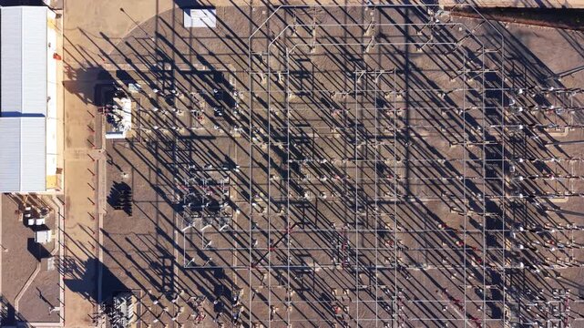 Overhead view of a large electrical substation with metal framework, power lines, transformers, and long shadows cast across a gravel yard beside an industrial building