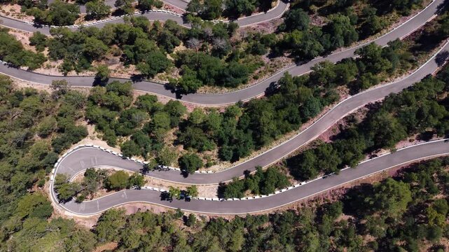 Overhead view of a winding serpentine mountain road with sharp hairpin bends cutting through dense green forest and dry scrubland terrain