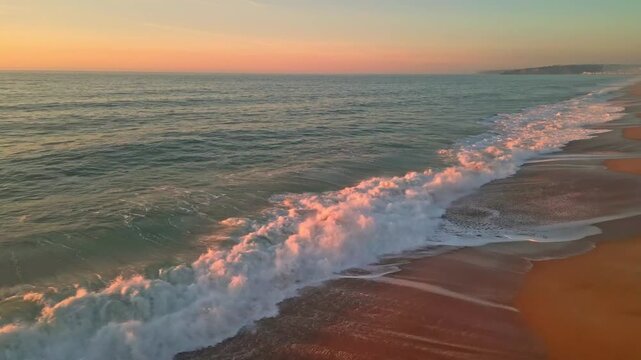 Cinematic aerial view of waves crashing at Praia do Salgado in Nazar&eacute;, Portugal. Beautiful golden hour light illuminates the Atlantic coast and sandy beach during a calm sunset