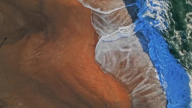 Stunning top-down slow-mo aerial video of Praia do Salgado in Nazare, Portugal. Cinematic view of Atlantic Ocean wave crashing against the golden beach sand with beautiful white foam patterns