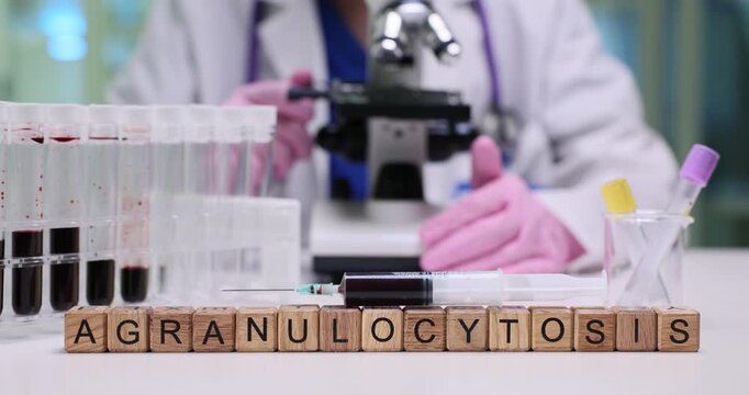 Agranulocytosis word spelled with wooden blocks and patients blood samples in lab. Female assistant looks in microscope working in medical center