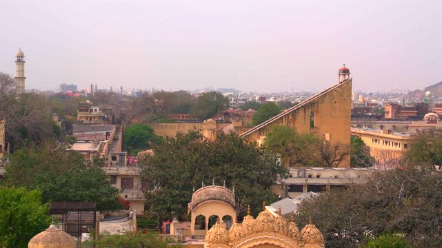 A wide static shot of the Jantar Mantar observatory skyline in Jaipur, featuring the massive Vrihat Samrat Yantra sundial and the historic Isar Lat tower in the city background.