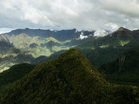 Aerial view of PR17 Pinaculo e Folhadal hiking trail along a lush green mountain ridge with a hiker in a yellow jacket under a cloudy sky in Serra de Agua, Madeira, Portugal.
