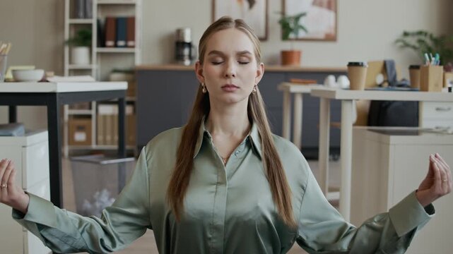Dolly in shot of young Caucasian woman meditating with eyes closed on yoga mat in modern office, practicing stress relief and preventing burnout