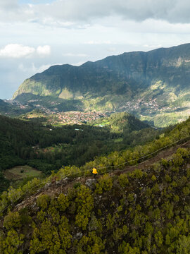 Aerial view of the PR17 Pinaculo e Folhadal hiking trail with a person in a yellow jacket overlooking the lush valley and mountains in Serra de Agua, Madeira, Portugal.