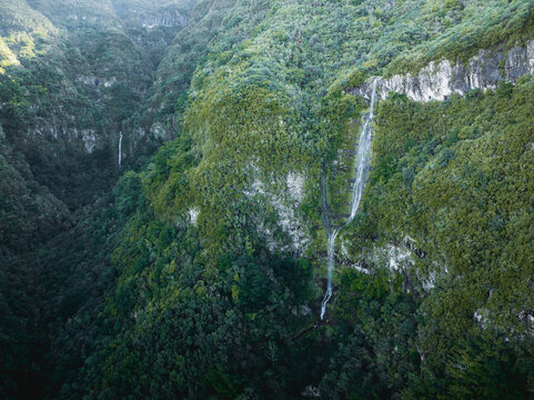 Aerial view of PR17 Pinaculo e Folhadal waterfall cascading down a steep rocky cliff surrounded by dense green forest and mountains in Serra de Agua, Madeira, Portugal.