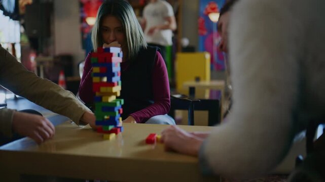 Young students gather around a table, intensely playing a game of Jenga. The group bursts into laughter as the colorful block tower collapses.
