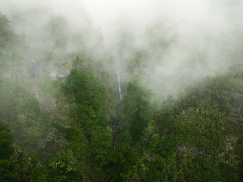 Aerial view of PR17 Pinaculo e Folhadal hiking trail area with waterfalls cascading down steep cliffs through dense green forest and thick mist in Serra de Agua, Madeira, Portugal.