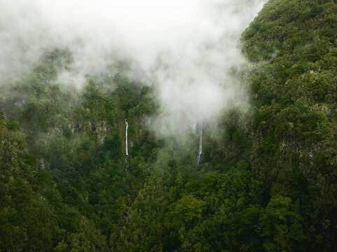 Aerial view of the PR17 Pinaculo e Folhadal trail featuring waterfalls cascading down lush green mountain slopes shrouded in thick white mist in Serra de Agua, Madeira, Portugal.
