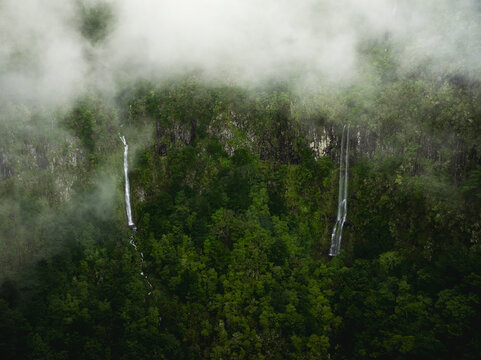 Aerial view of PR17 Pinaculo e Folhadal area with twin waterfalls cascading down cliffs into a dense green forest shrouded in thick mist Serra de Agua, Madeira, Portugal.