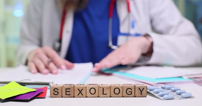 Word Sexology on wooden cubes near pills and condoms on doctor desk. Female medic prepares patient records for regular appointment in clinic