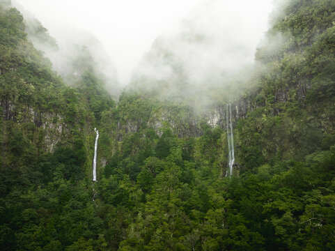 Aerial view of two waterfalls cascading down lush green cliffs partially covered by thick white fog in Serra de Agua, Madeira, Portugal.