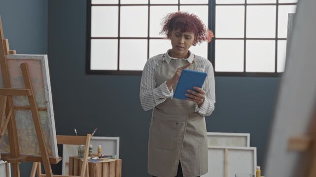 Woman painter taps a blue tablet while studying reference images in an art studio surrounded by easels, canvases, brushes, paint tubes and a window; creative concentration.