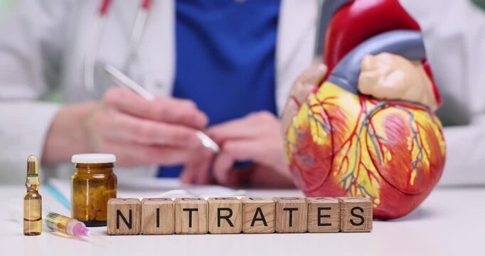 Wooden blocks spell word Nitrates near heart model and tablets. Doctor writes cardiology notes reviewing vasodilation use for chest pain care