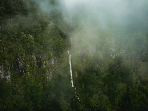 Aerial view of the PR17 Pinaculo e Folhadal waterfall cascading down a steep green cliff surrounded by dense forest and mist in Serra de Agua, Madeira, Portugal.