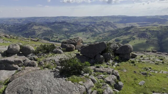 Drone flies over stacked rocks on edge of cliff overlooking green valley on a sunny day at Sibebe Rock near Mbabane, Eswatini
