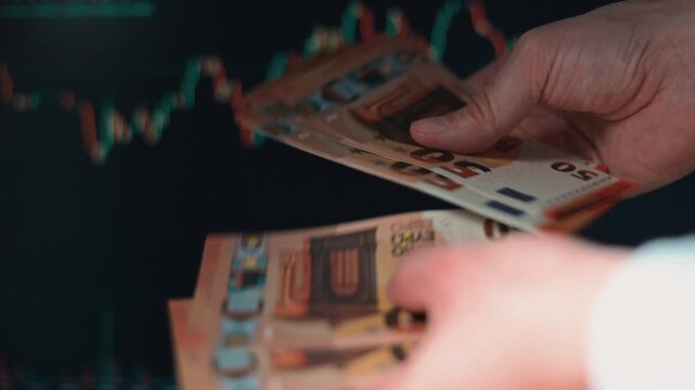 A person sorts and counts euro cash while looking at financial data on a screen. The setting suggests a focus on finance and financial management during a work session