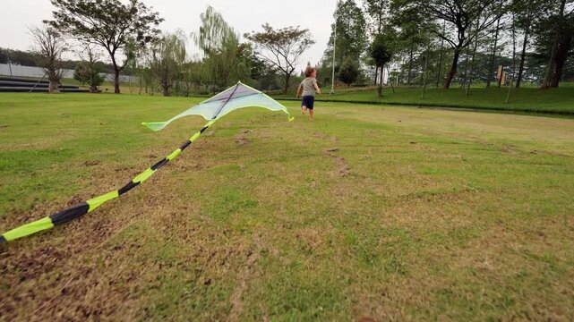 Little boy glances back at his kite lying on ground, then turns and runs forward, pulling kite on short string. Kite quickly takes off into air. In slow motion, camera follows from behind