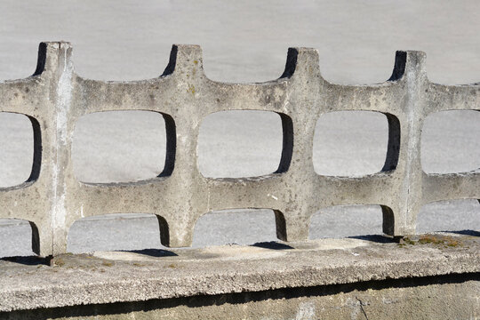 A close up shot of a weathered decorative concrete breeze block fence with geometric cutouts standing on a stone base under bright sunlight