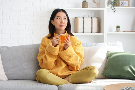 Beautiful young Asian woman with cup of tea sitting on sofa in living room