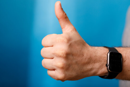 Close-up of a hand with a thumbs-up gesture, smart watch on wrist with blue background.