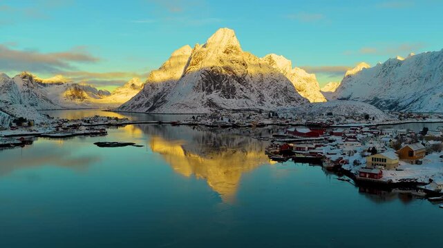 Cinematic aerial shot of Reine village in Lofoten, Norway, featuring snowcapped mountains reflecting in calm fjord waters during golden hour.