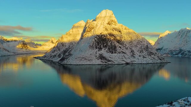 A cinematic aerial shot of snowcapped peaks reflecting in the calm blue waters of a fjord in Lofoten, Norway, during a vibrant winter sunset.
