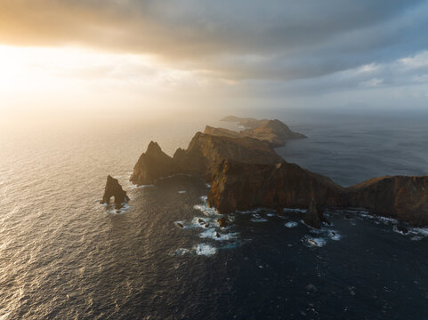Aerial view of Ponta de Sao Lourenco with rugged volcanic cliffs and crashing waves under a golden sunset sky in Canical, Madeira, Portugal.