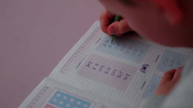 Child practicing math exercises with green pencil, writing on graph paper notebook, showing visible calculations and colorful illustrations in a bright indoor setting