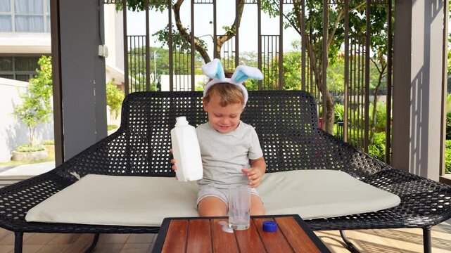 Cheerful talking toddler pours milk from white plastic bottle into glass. He pours gently at first, then more vigorously, spilling bit on table. Unbothered by mess, he smiles brightly