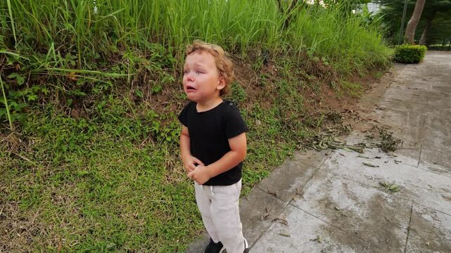 Frightened toddler stands on sidewalk, crying with tearful eyes and open mouth. Young child is visibly upset, overwhelmed by fear and distress. His face is tear-streaked, and he looks very nervous.