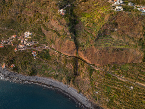 Aerial view of a waterfall cascading down a steep cliff onto a coastal road, with terraced agriculture and white houses overlooking the Atlantic Ocean in Ponta do Sol, Madeira, Portugal.