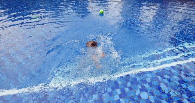 Brave cheerful little boy fearlessly falls backward into swimming pool. Slow-motion captures his joyful face and big splash he creates. Funny toddler enjoys water play on hot day